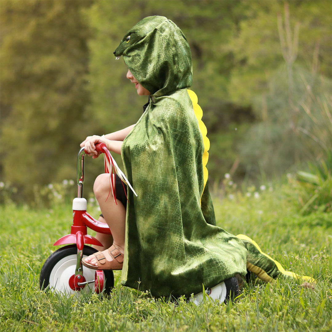 Een kind zit op een rode driewieler in het gras, gekleed in de groene dino T-Rex verkleedcape.