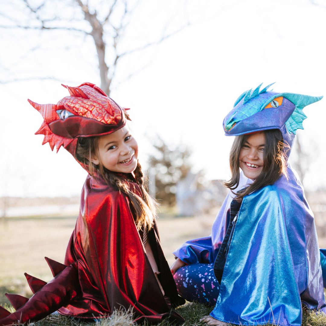 Twee lachende kinderen, waarvan één in een great pretenders rode metallic draken verkleedcape, zitten buiten in het gras.