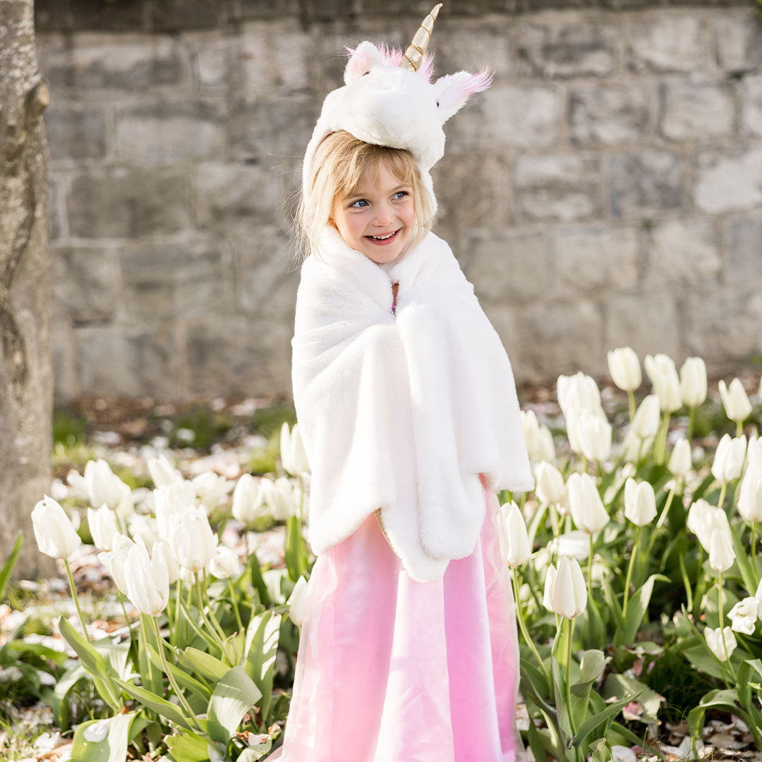 Een lachend kind draagt de witte eenhoorn verkleedcape en een roze jurk, staand tussen witte tulpen.
