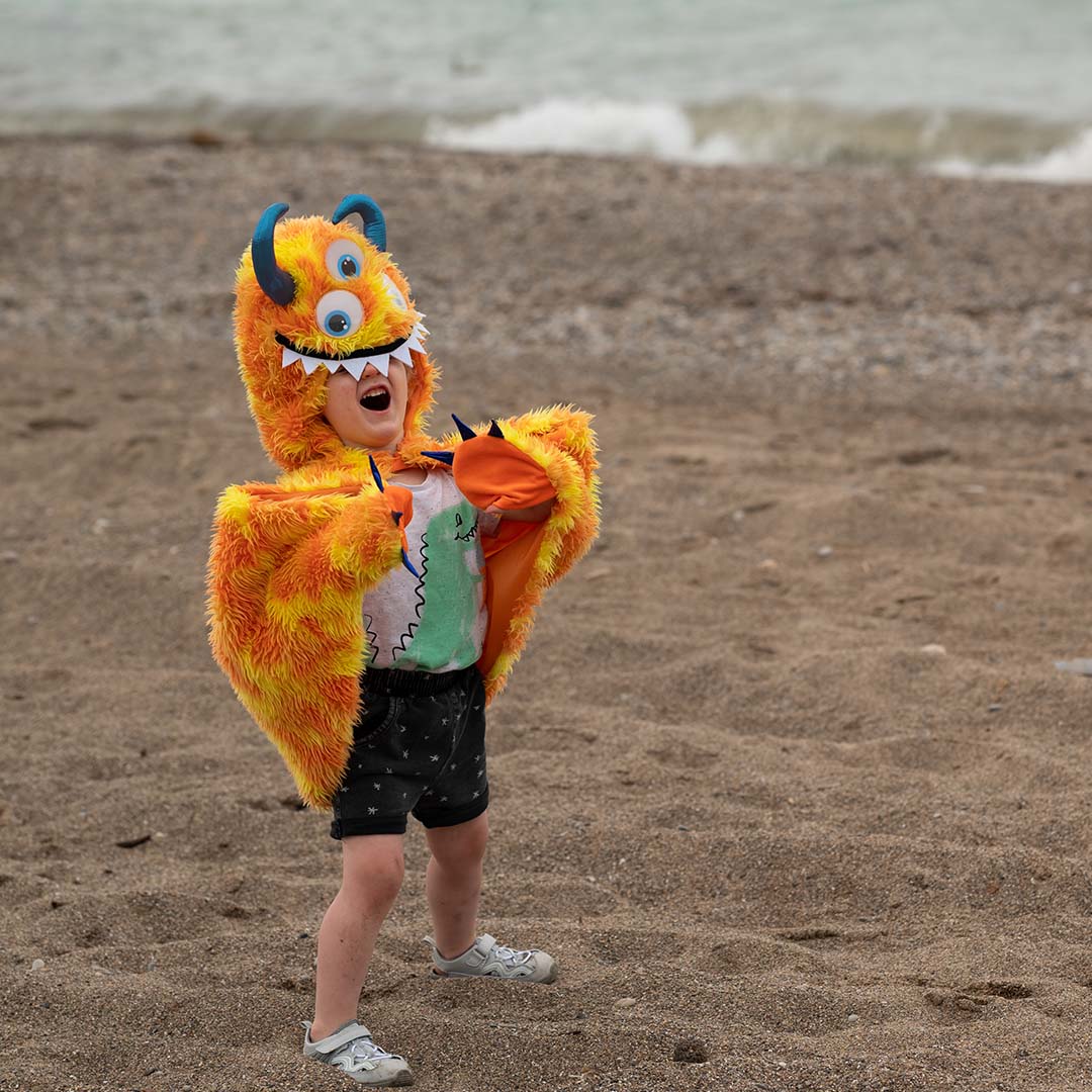 Een kind draagt de oranje en gele monster verkleedcape op een zandstrand.