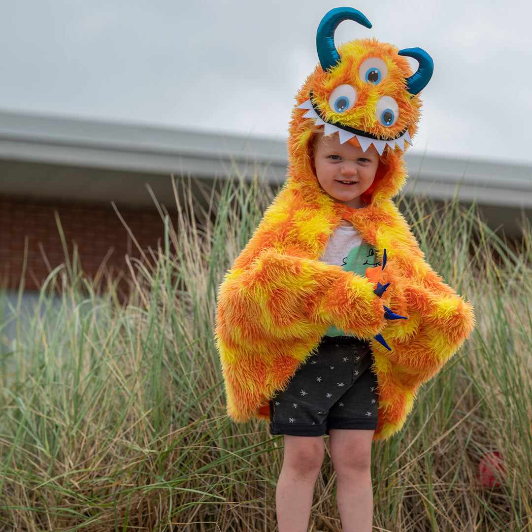 Een lachend kind draagt de oranje en gele monster verkleedcape voor een achtergrond van duingras.