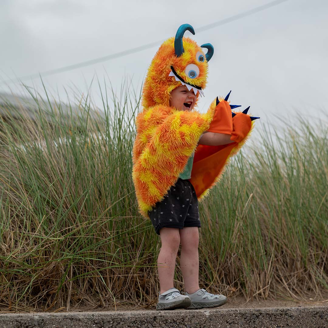 Een kind lacht terwijl het de oranje en gele monster verkleedcape draagt met gespreide armen.