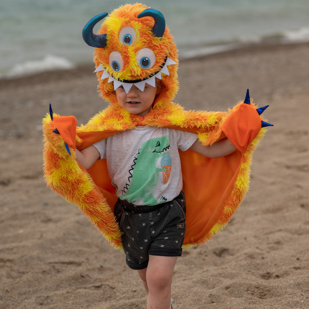 Een kind draagt de oranje en gele monster verkleedcape op een zandstrand met de zee op de achtergrond.