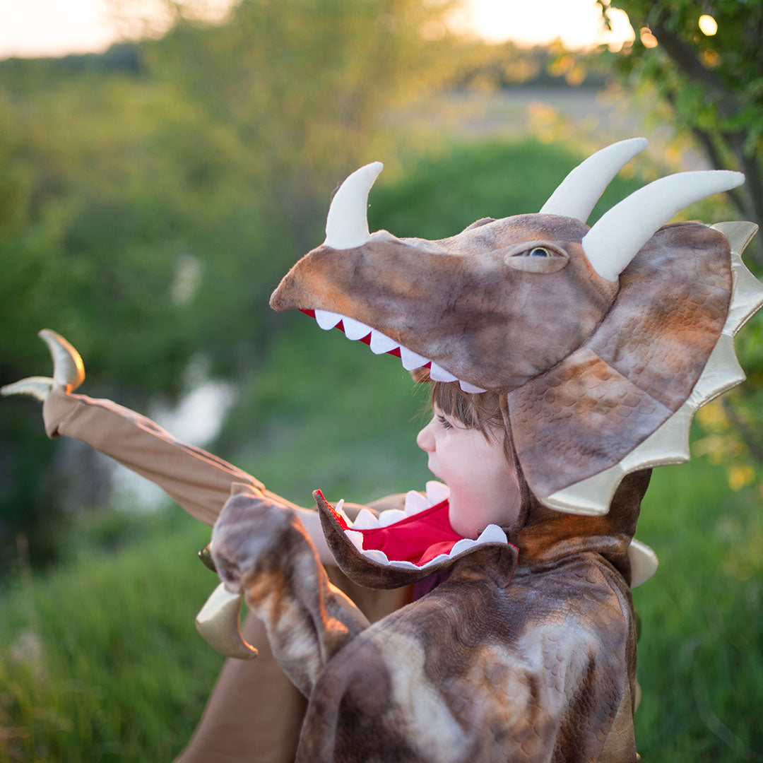 Een kind draagt de triceratops verkleedcape en kijkt omhoog in een groene buitenomgeving.