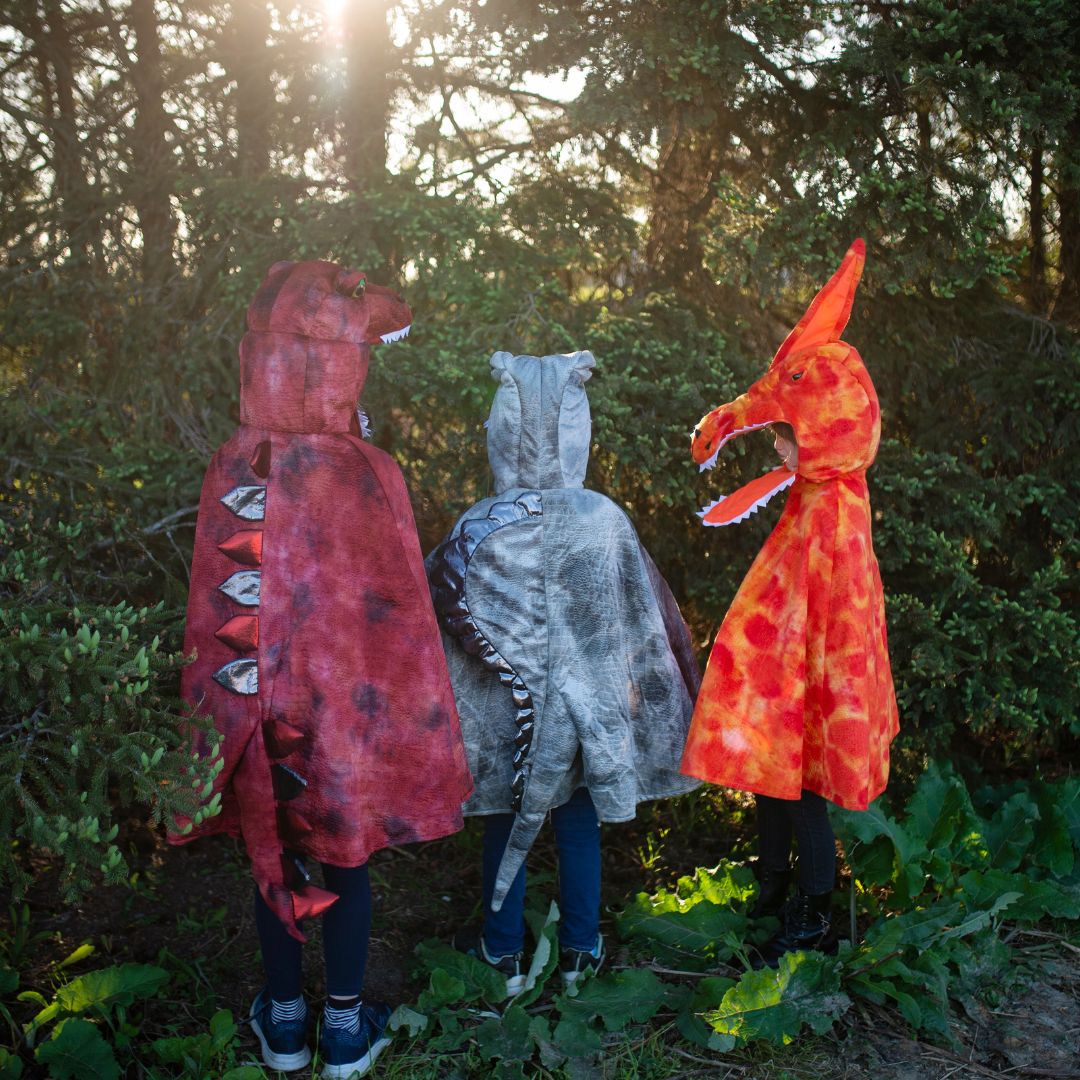 Drie kinderen in dinosaurus verkleedcapes, waaronder een rode T-Rex, van achteren gezien in een bos.