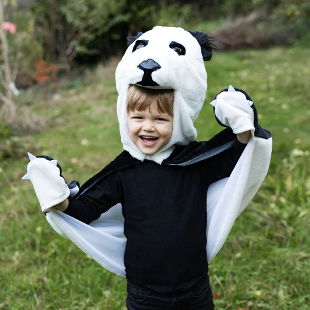 Lachende peuter draagt de Great Pretenders panda verkleedcape met opgestoken armen in het gras.