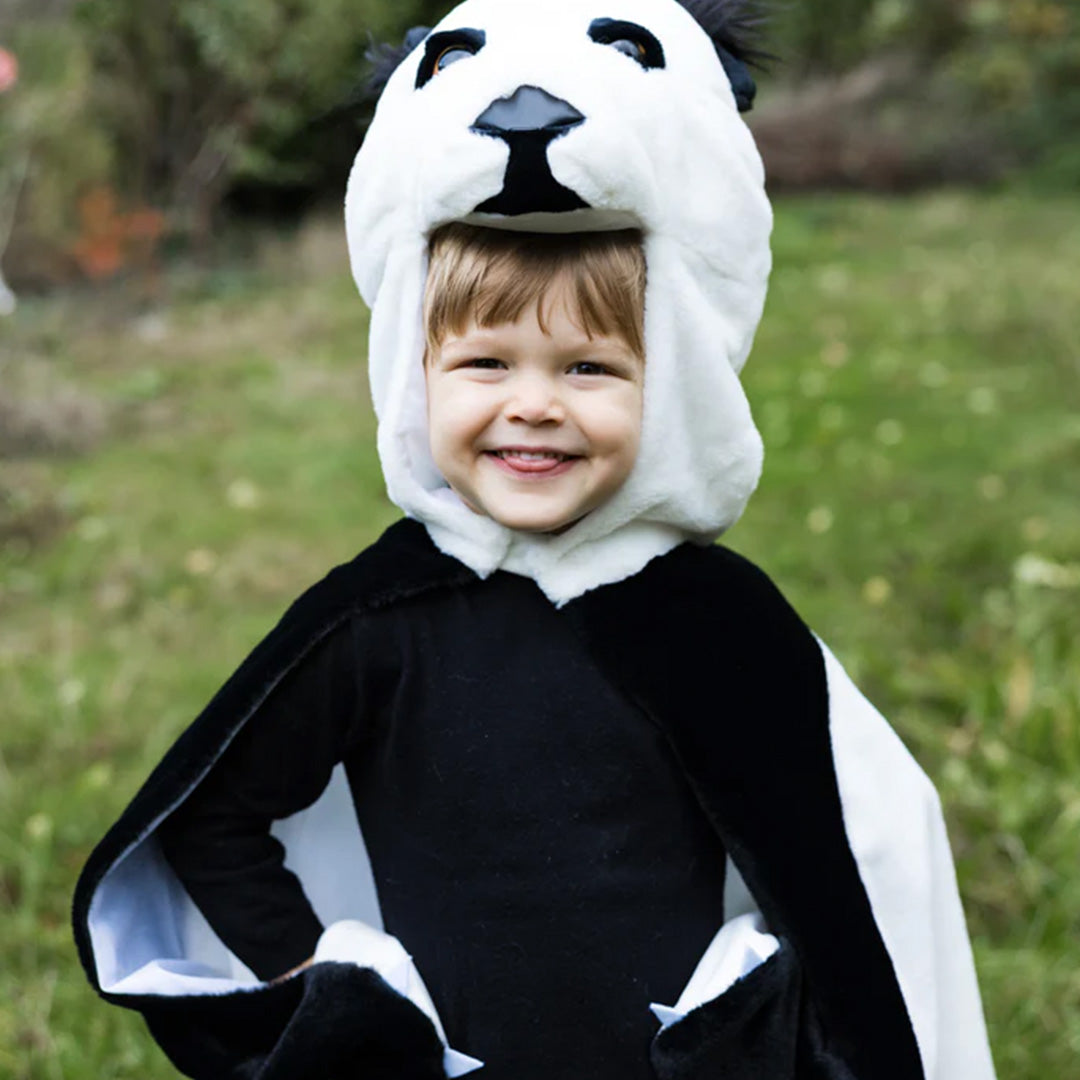 Lachende peuter draagt de Great Pretenders panda verkleedcape met de handen in de zij in het gras.