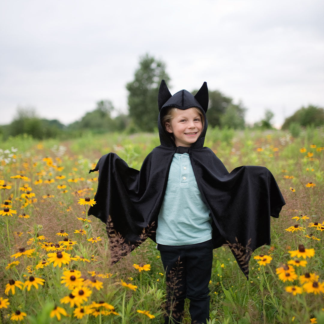 Een lachend kind draagt de zwarte verkleedcape vleermuis met gespreide vleugels in een veld met gele bloemen.