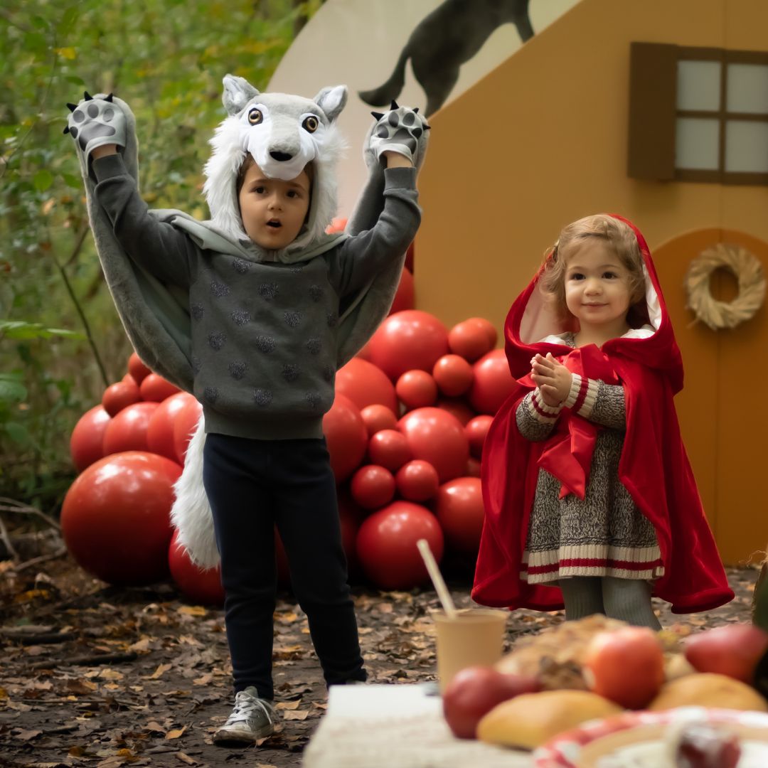 Twee kinderen in een bosrijke omgeving, één draagt de Great Pretenders grijze wolf verkleedcape met de handen omhoog en de ander een roodkapje cape.