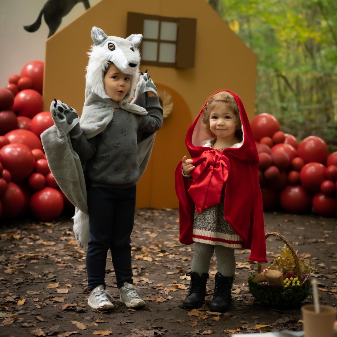Twee kinderen in een bosrijke omgeving, één draagt de Great Pretenders grijze wolf verkleedcape met de handen omhoog en de ander een roodkapje cape.