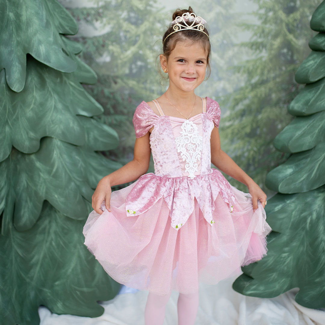 Lachend meisje draagt de dusty rose prinsessen verkleedjurk met een tiara, staand voor groene decoratieve bomen.