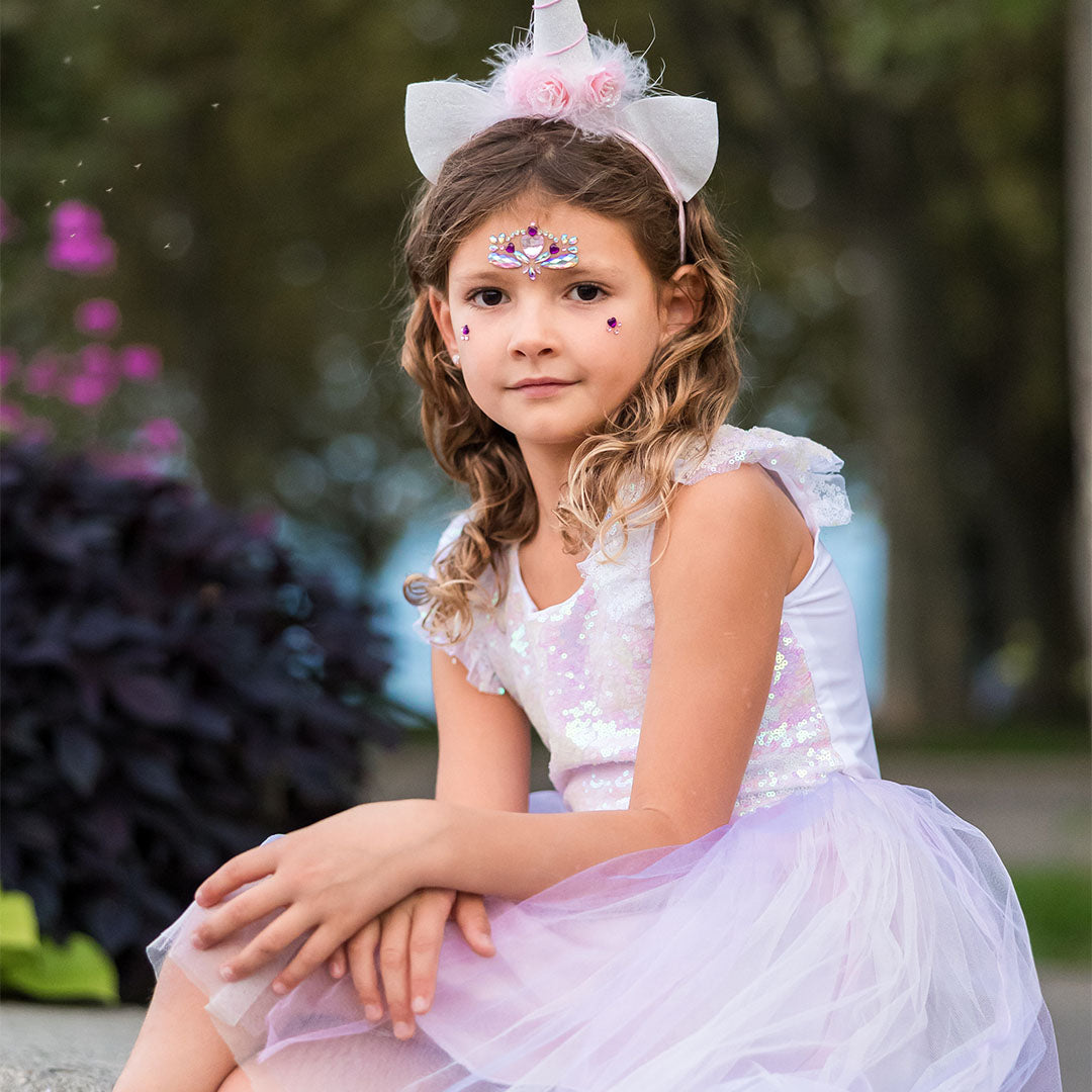 Meisje in de eenhoorn verkleedjurk met diadeem en glitter make-up zit buiten.