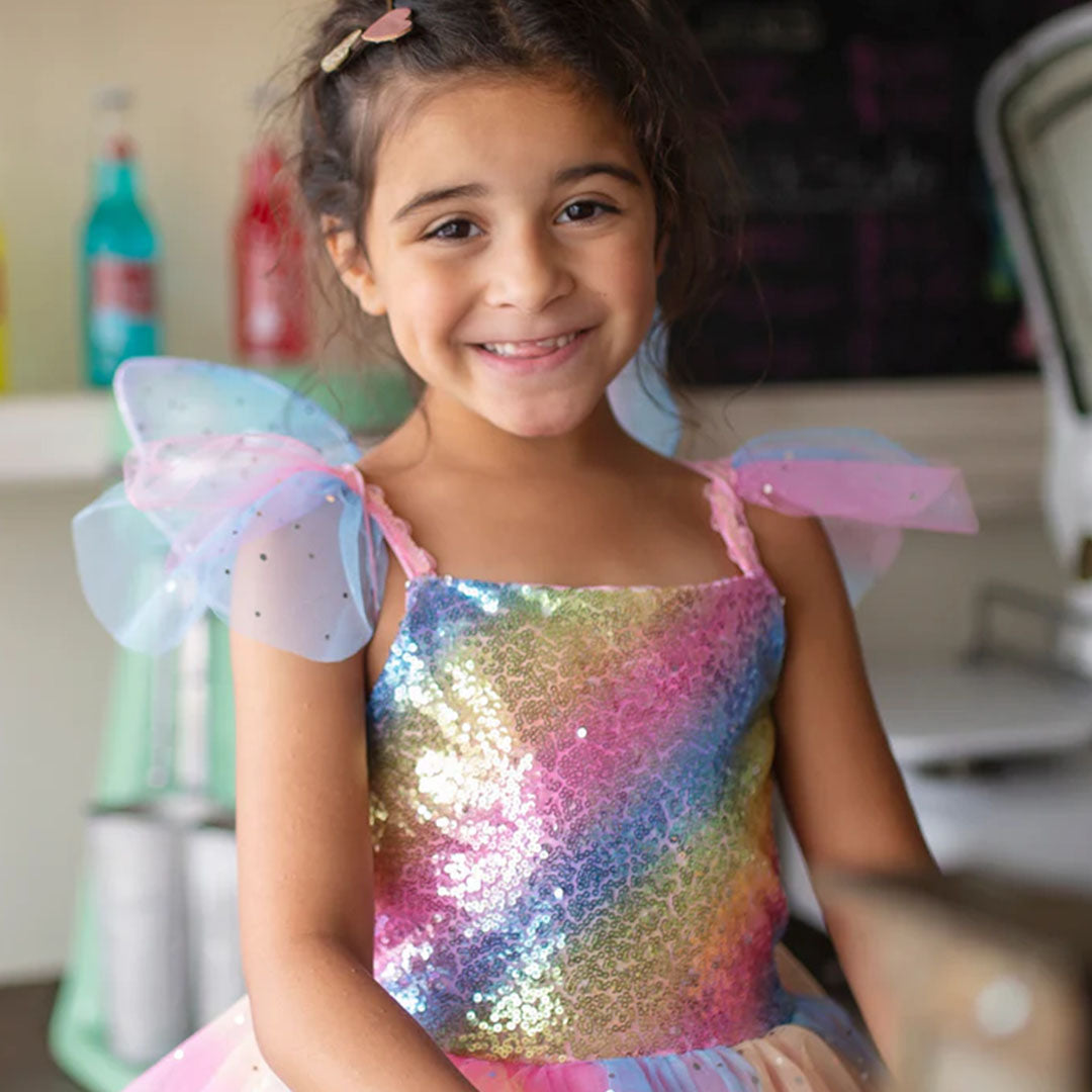 Een lachend meisje draagt de Great Pretenders regenboog elfjes verkleedjurk met pailletten en kleine vleugels.