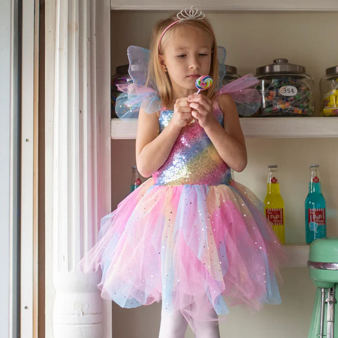 Een meisje draagt de Great Pretenders regenboog elfjes verkleedjurk met pailletten en kleine vleugels, terwijl ze een lolly vasthoudt.