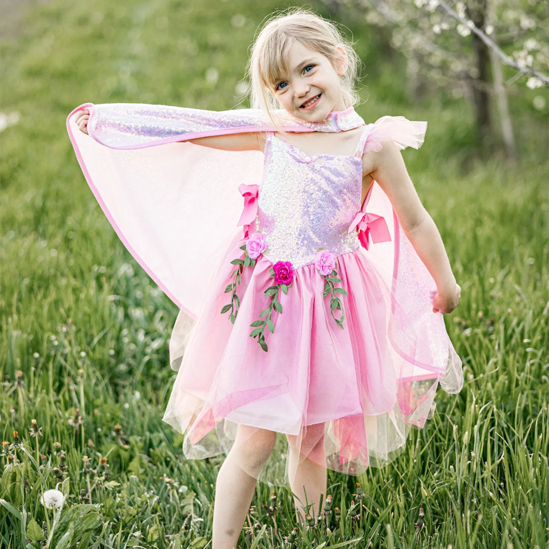 Een lachend meisje draagt de Great Pretenders roze sprookjes verkleedjurk met cape in een groen veld.
