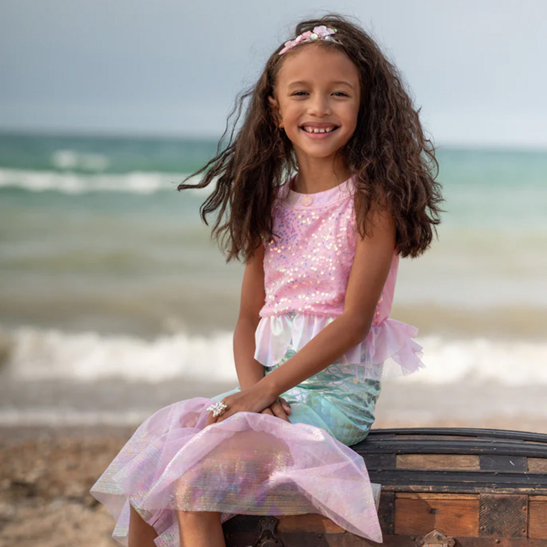 Een lachend meisje in de zeemeermin verkleedjurk zit op een schatkist aan het strand.