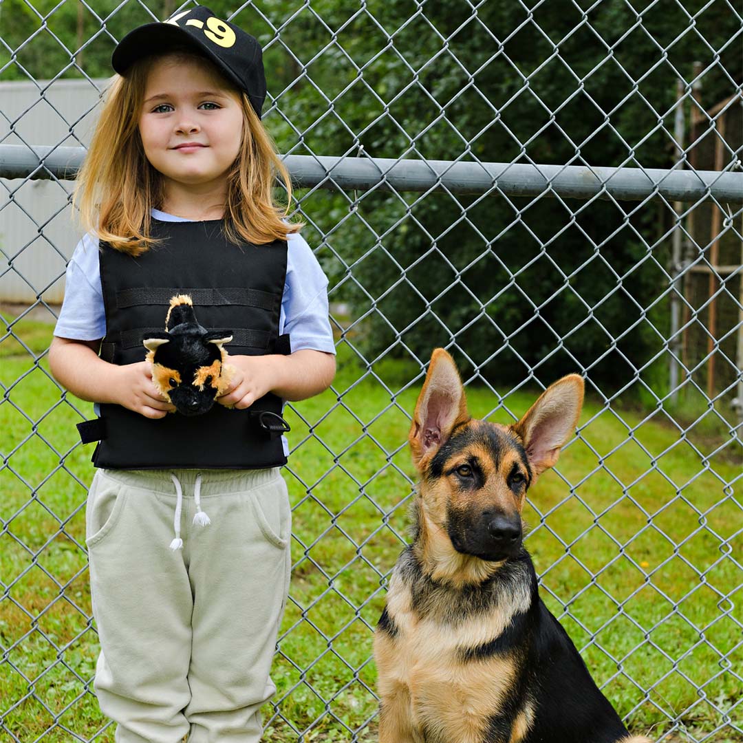 Meisje in K-9 politieagent verkleedkleding met pluche hond naast een echte Duitse herder bij een hek.