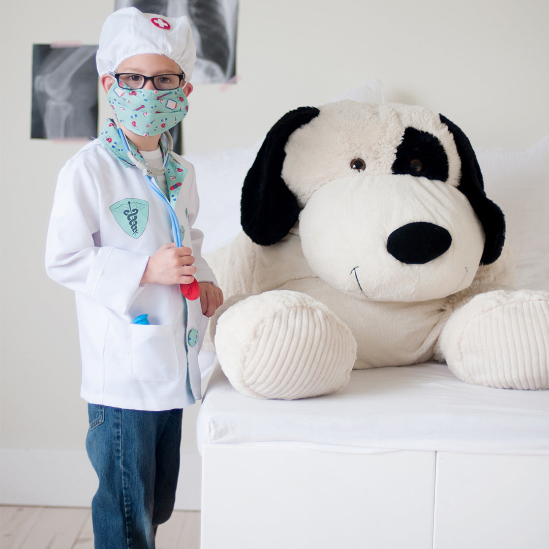 Een kind in Great Pretenders dokter verkleedkleding met stethoscoop onderzoekt een grote knuffelhond op bed.