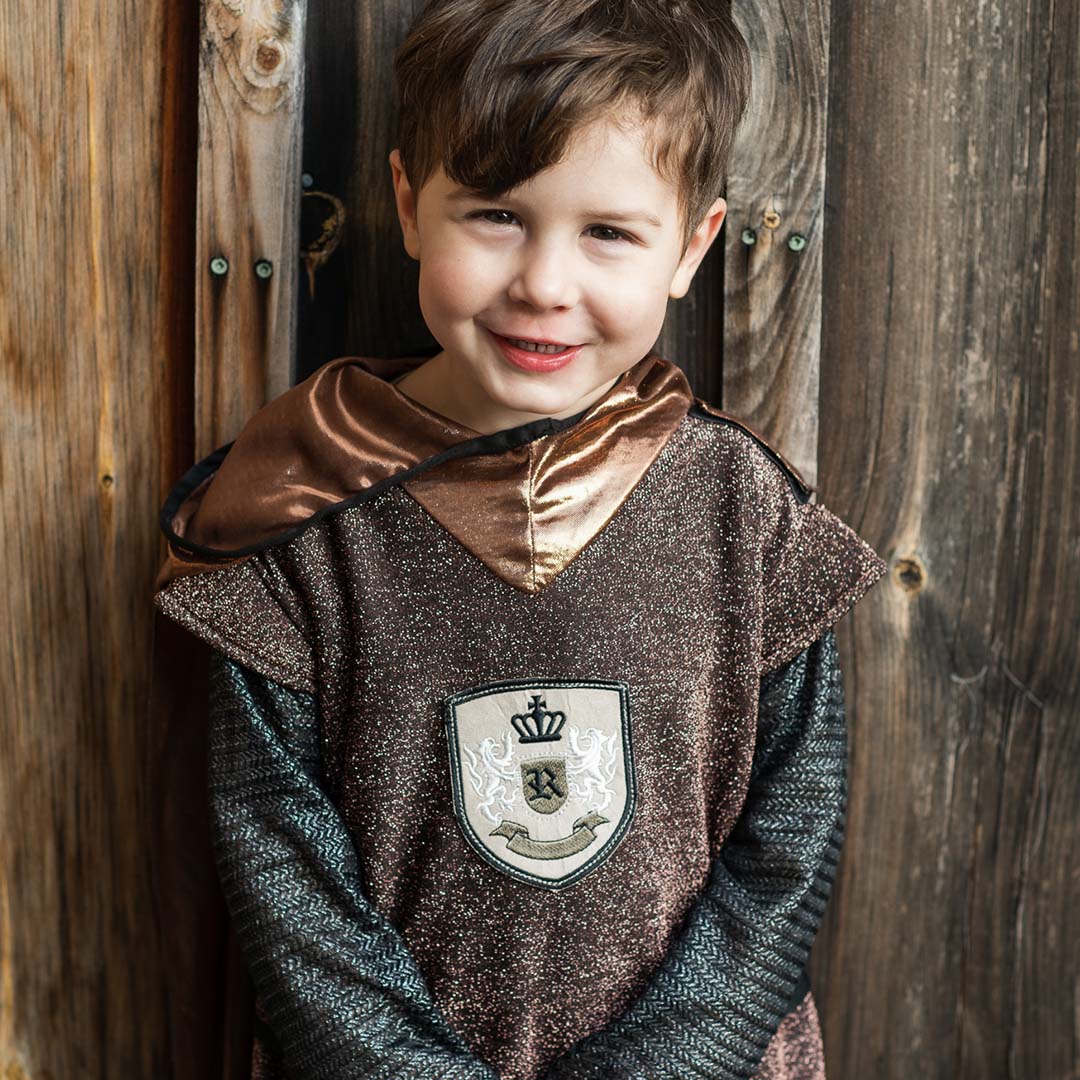 Lachende jongen draagt het Great Pretenders kinderkostuum ridder voor een houten schutting.