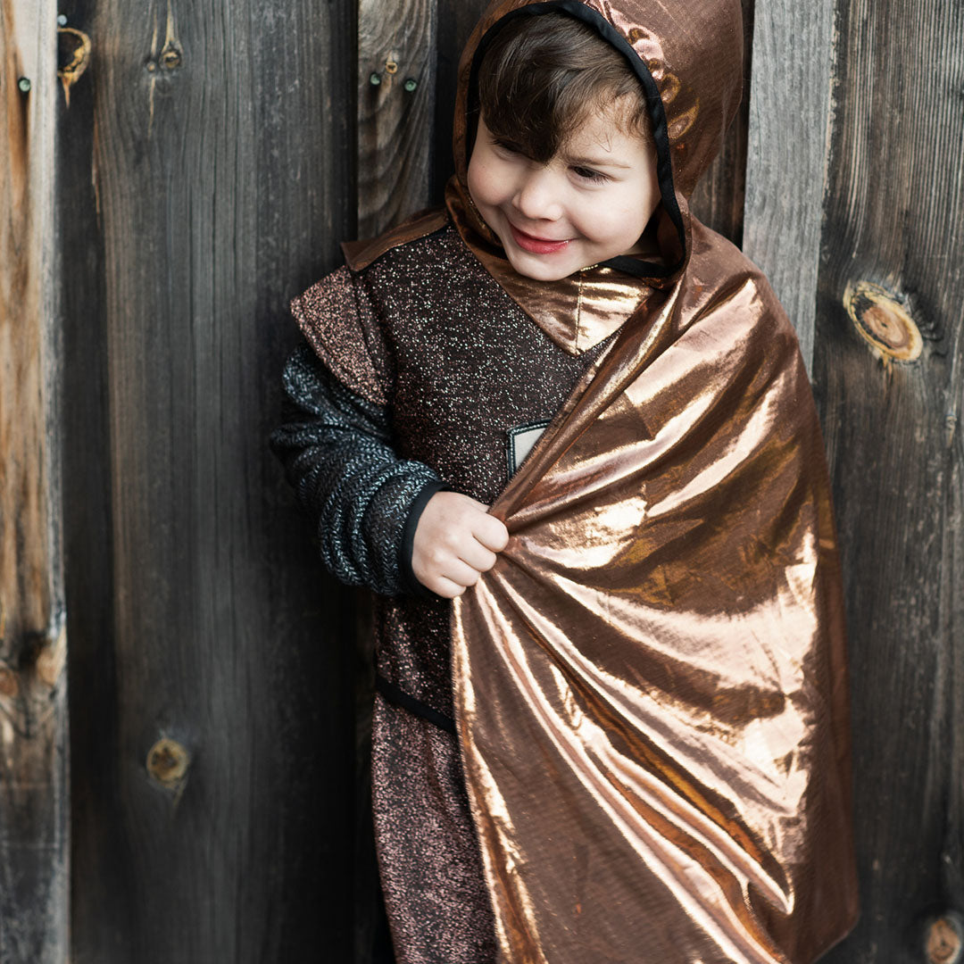 Jongen in het Great Pretenders kinderkostuum ridder lacht en houdt de bronzen cape vast.