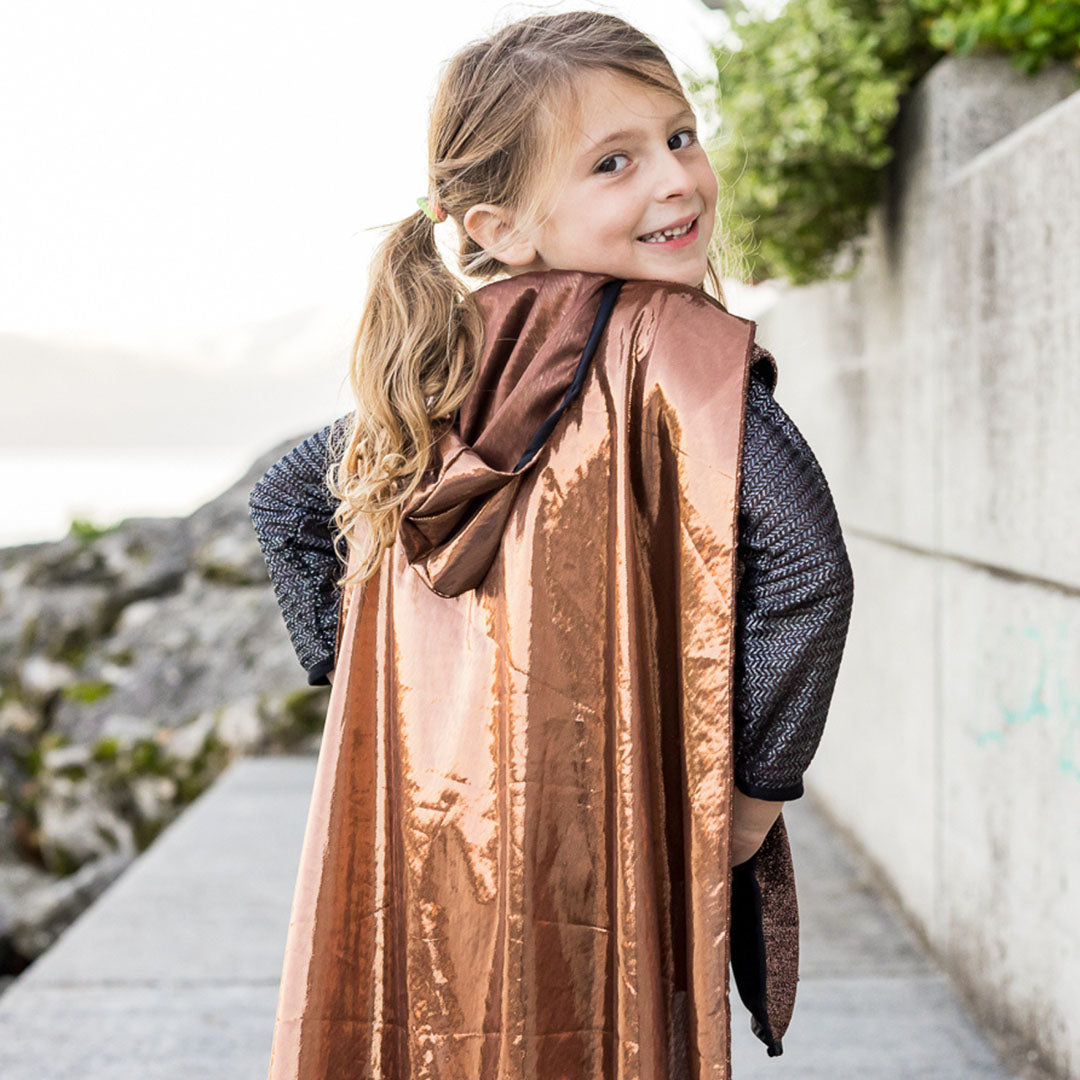 Meisje in het Great Pretenders kinderkostuum ridder kijkt lachend over haar schouder, met de bronzen cape op de rug.