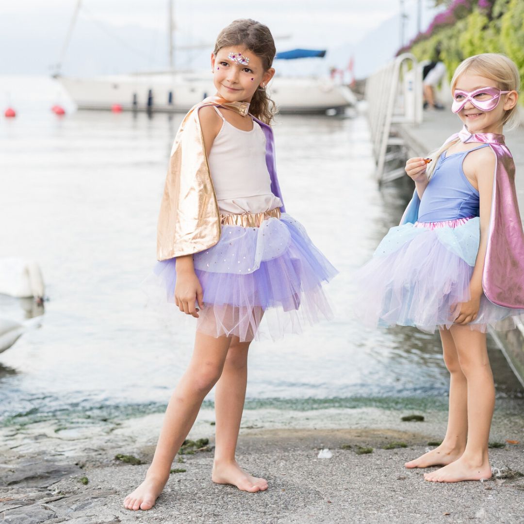 Twee meisjes in great pretenders superheld kostuumsets, één met gouden cape en paarse rok, staan bij het water.