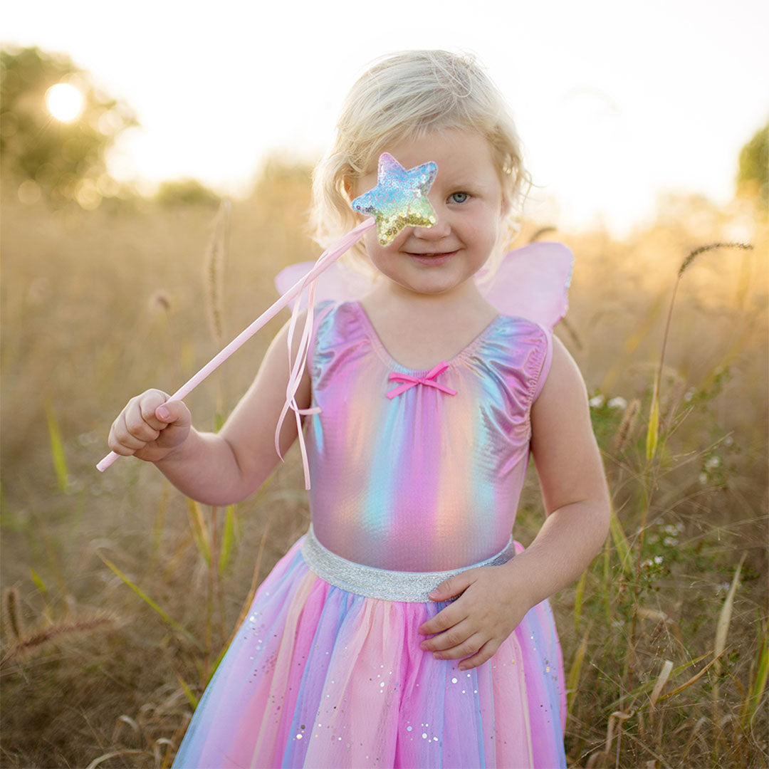 Een lachend kind draagt de great pretenders regenboog verkleedset en houdt de toverstaf voor haar oog in een veld.