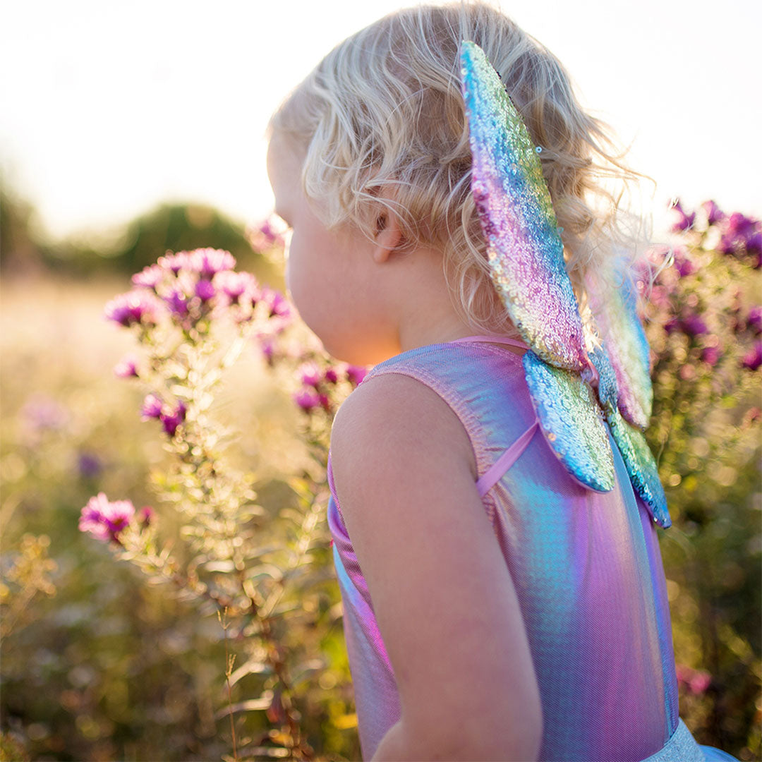 Een kind draagt de great pretenders regenboog verkleedset met vleugels en kijkt naar paarse bloemen in een veld.