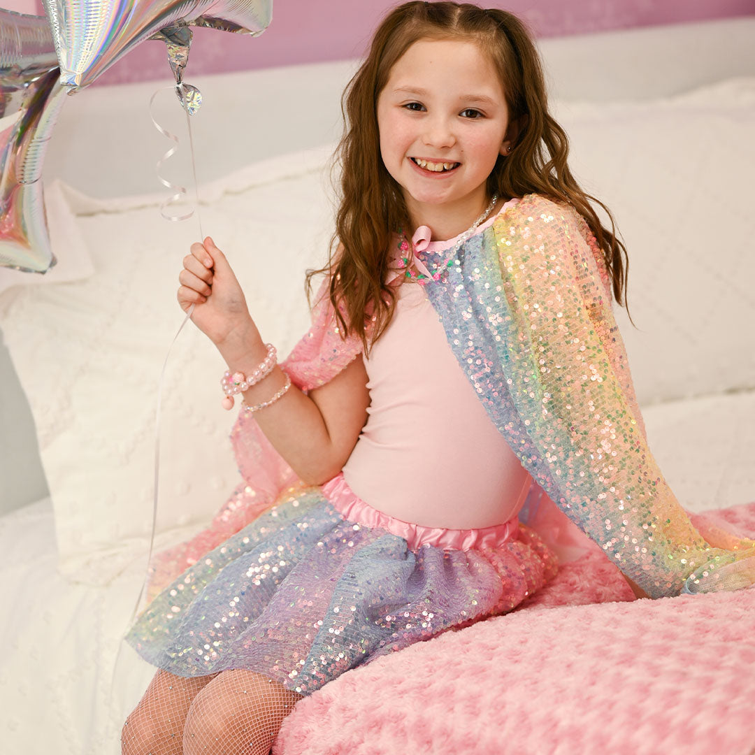 Een lachend kind draagt de great pretenders verkleedrok met regenboog pailletten en een bijpassende cape, zittend op een bed.