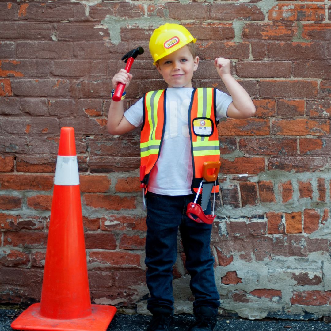 Jongen van 5-6 jaar in bouwvakker verkleedset met gele helm en oranje vest, staand bij een oranje pion.