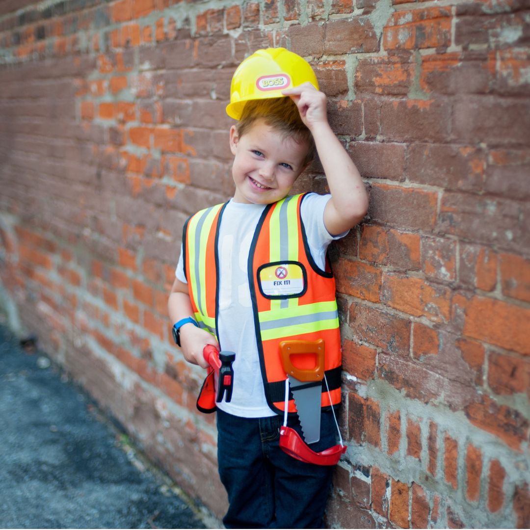 Lachende jongen van 5-6 jaar in bouwvakker verkleedset met gele helm en oranje vest, leunend tegen een bakstenen muur.
