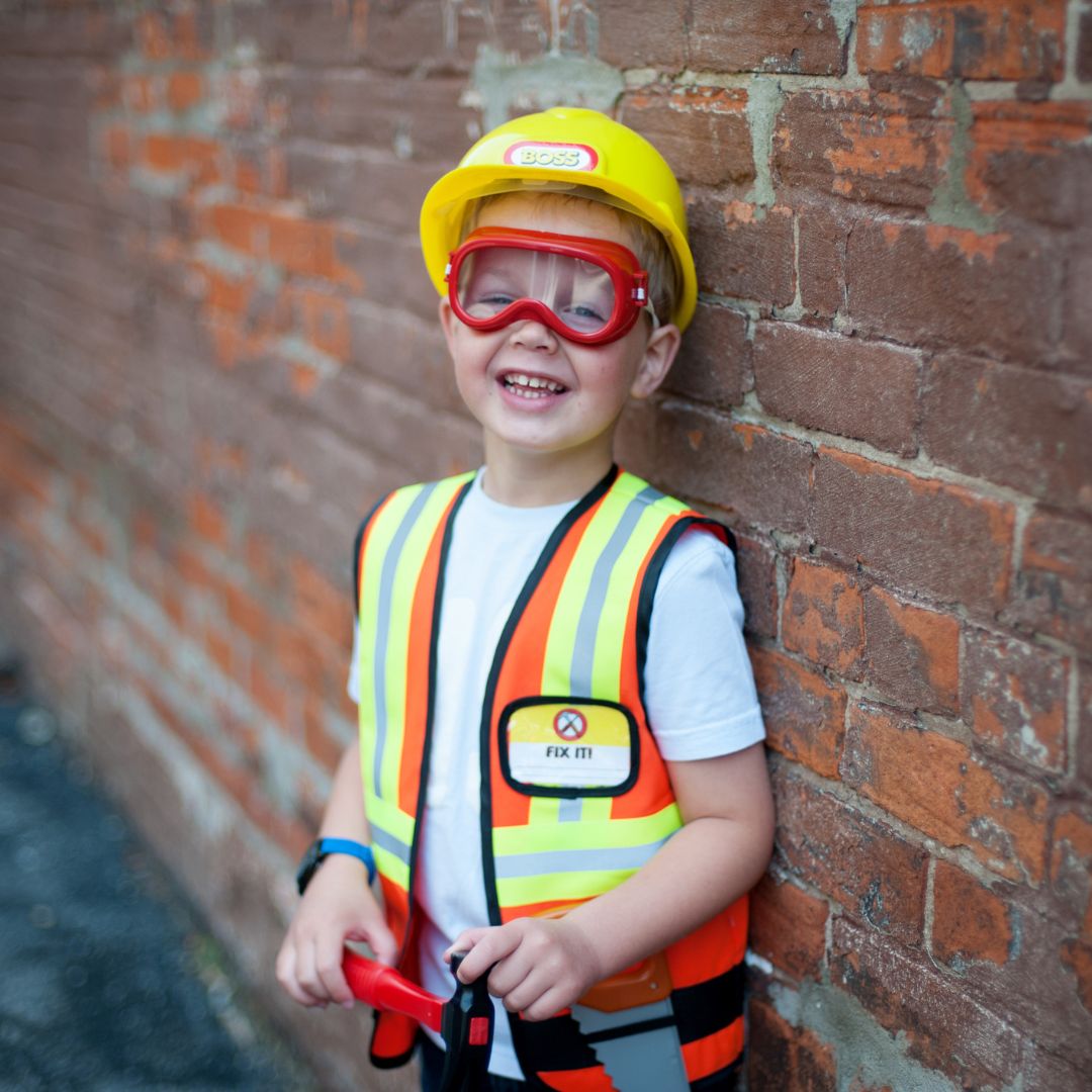 Lachende jongen van 5-6 jaar in bouwvakker verkleedset met gele helm, oranje vest en rode veiligheidsbril.