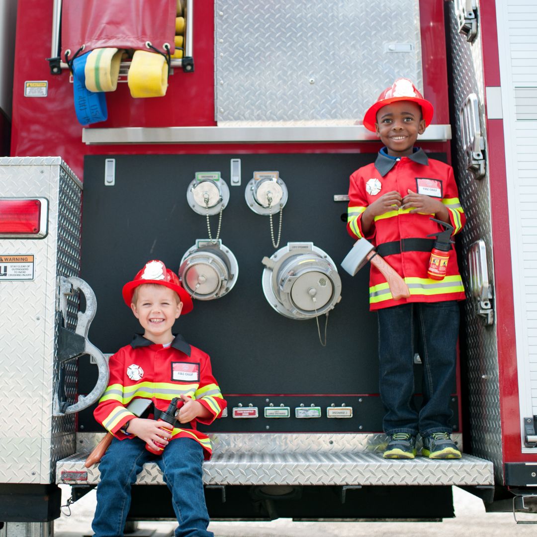 Twee jongens in brandweerman verkleedkleding staan bij de achterkant van een brandweerwagen.