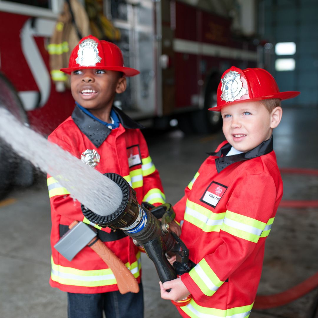 Twee jongens in Great Pretenders brandweerman verkleedsets spuiten water met een brandweerslang.