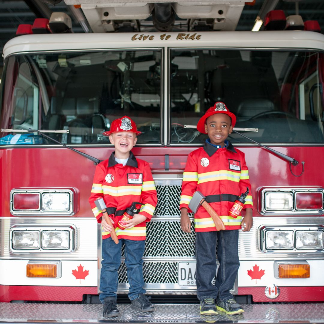 Twee lachende jongens in Great Pretenders brandweerman verkleedsets staan voor een rode brandweerwagen.