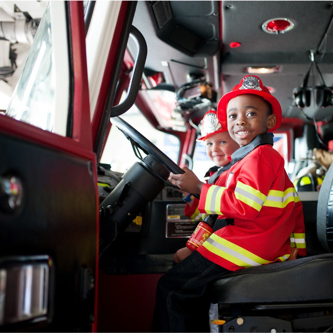 Een lachende jongen in een Great Pretenders brandweerman verkleedset zit achter het stuur van een brandweerwagen.