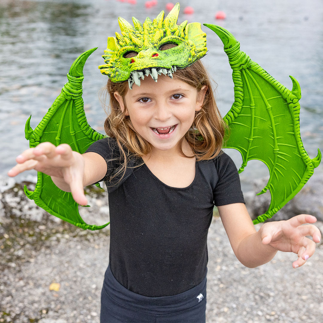 Lachend kind met Great Pretenders groene drakenvleugels en drakenmasker reikt naar voren.