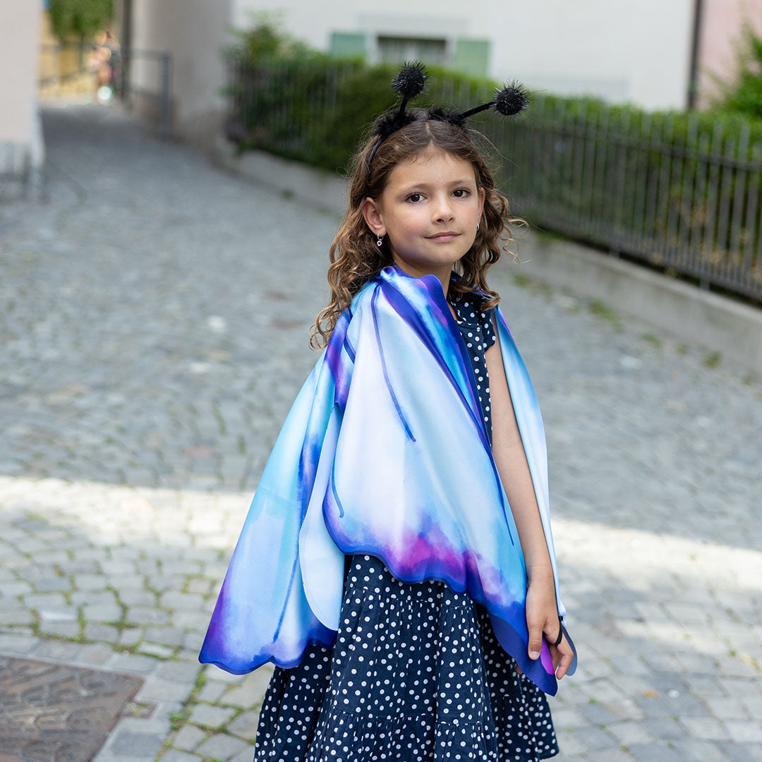 Een meisje draagt de blauw-paarse vlindervleugels en diadeem, staand op een straat met kinderkopjes.