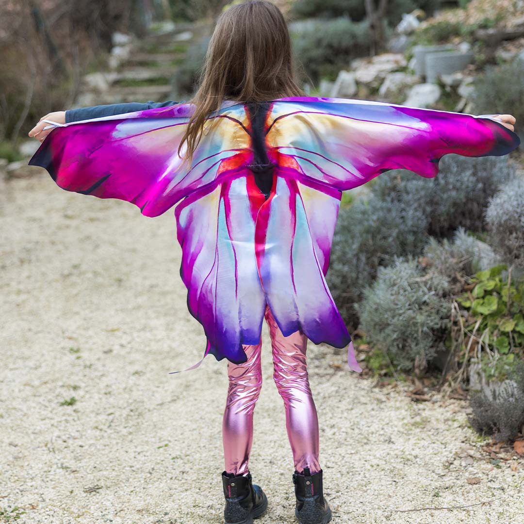 Kind van achteren met Great Pretenders vlinder vleugels en roze broek, met gespreide armen in een tuin.