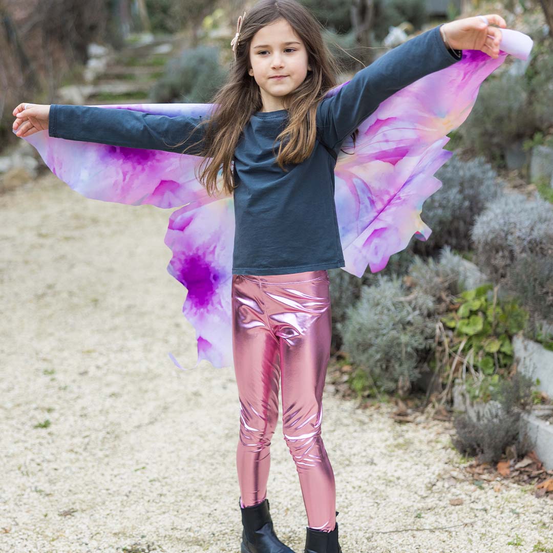 Kind met Great Pretenders vlinder vleugels en roze broek, met gespreide armen in een tuin.