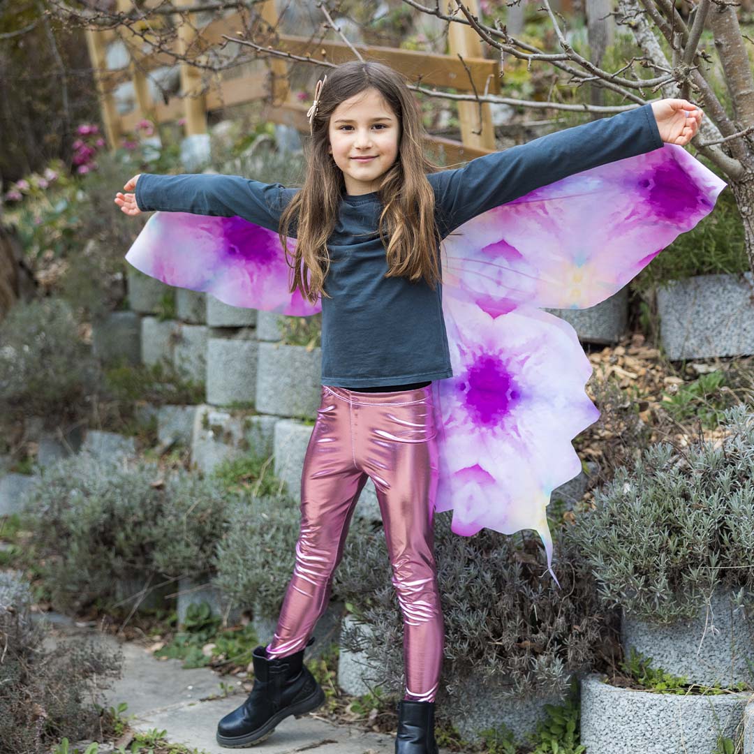 Lachend kind met Great Pretenders vlinder vleugels en roze broek, met gespreide armen in een tuin.