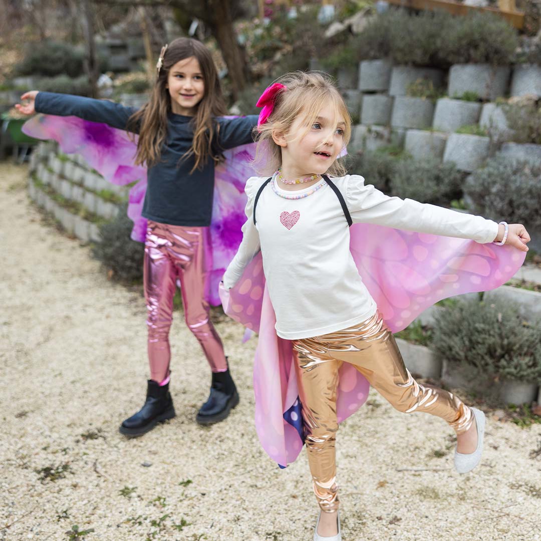 Twee kinderen met Great Pretenders vlinder vleugels en glimmende broeken in een tuin.