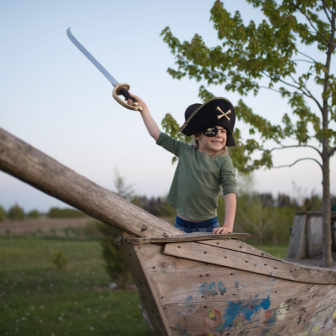 Kind met Great Pretenders piraten ooglapje en hoed, staand in een houten boot met een speelgoedzwaard.