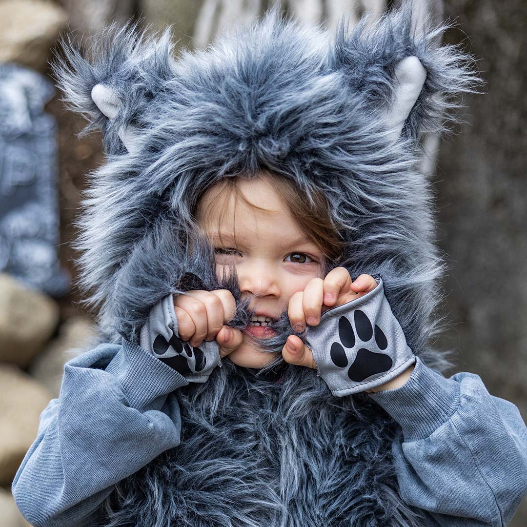 Close-up van een kind in de grijze wolf verkleedcape, dat speels de vacht rond het gezicht trekt met de pootmanchetten.