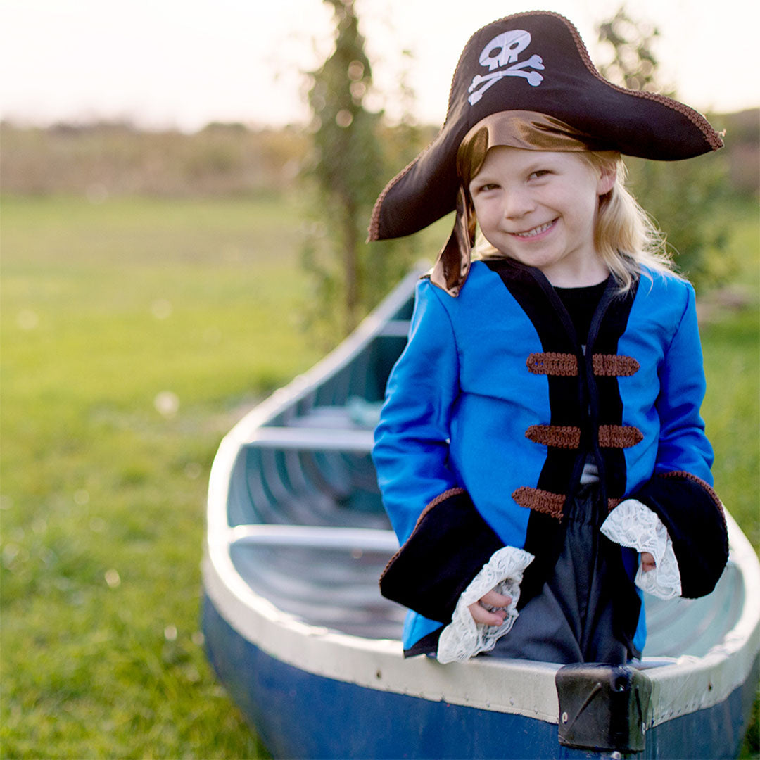 Een lachend kind in een blauw piratenkostuum met hoed zit in een kano in het gras.