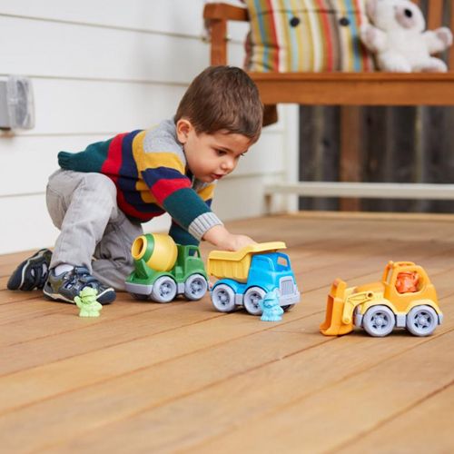 Een jong kind speelt op een houten vloer met een gele kiepwagen, een groene cementwagen en een oranje shovel.