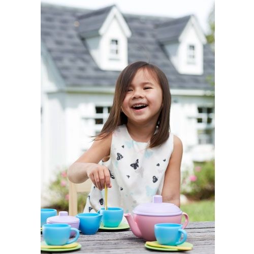 Een lachend meisje speelt buiten met het Green Toys theeservies aan een houten tafel, met een wit huis op de achtergrond.