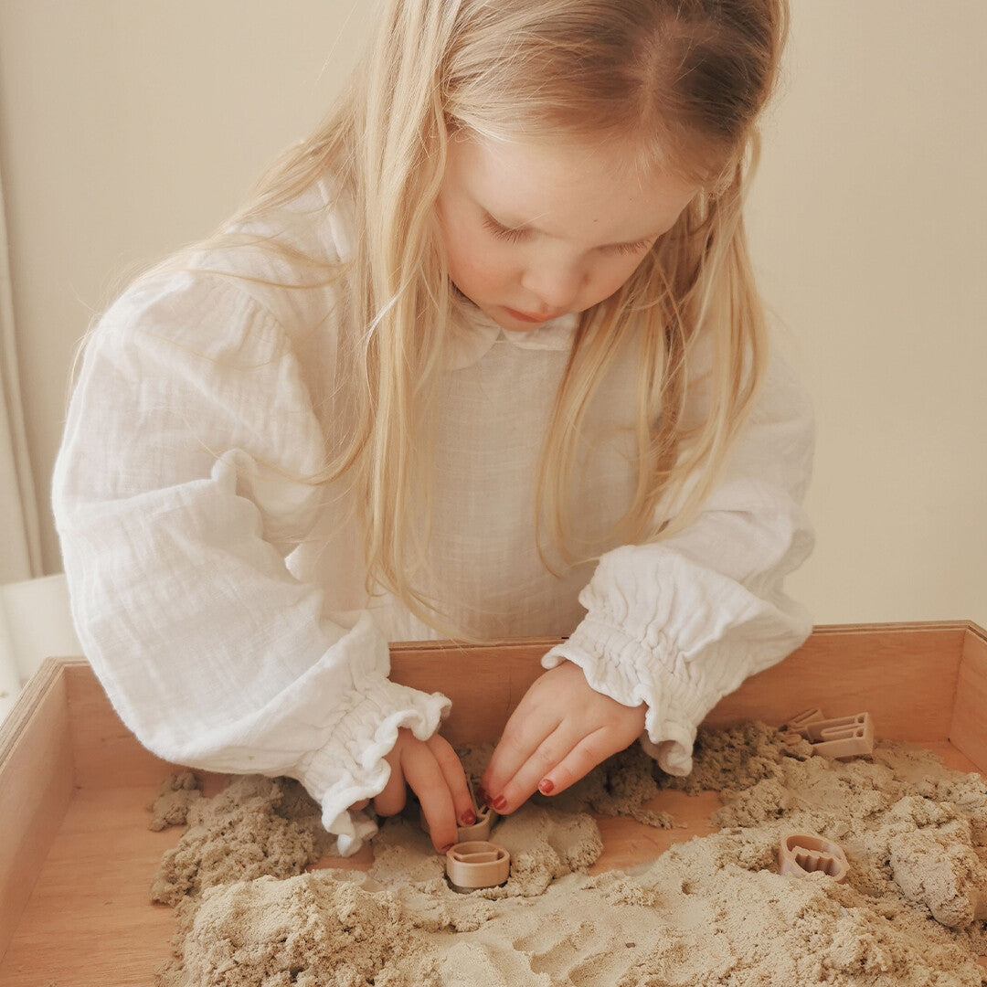 Een meisje met blond haar speelt met beige kinetisch zand en kleine vormpjes in een houten bak.