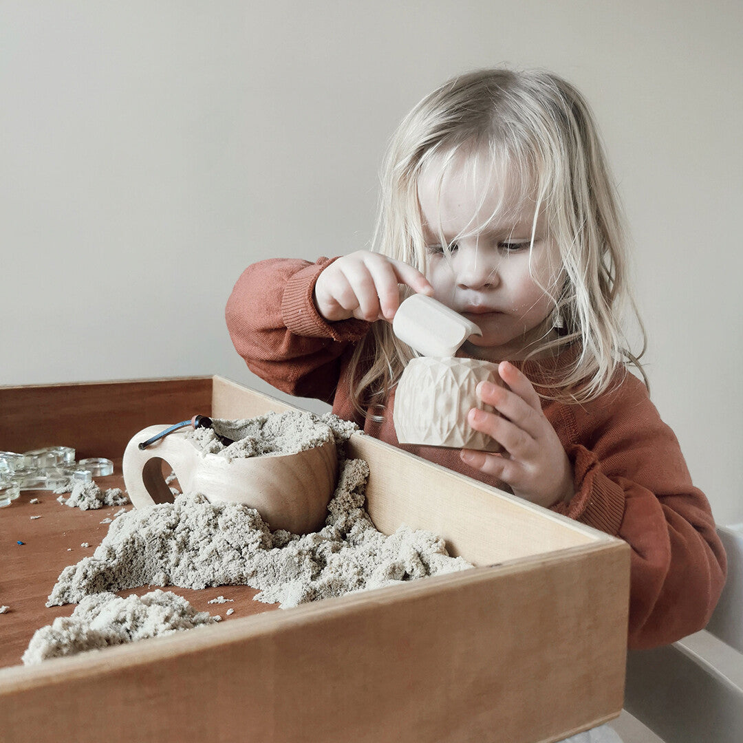 Een jong kind speelt met beige kinetisch zand en een schepje in een houten bak.