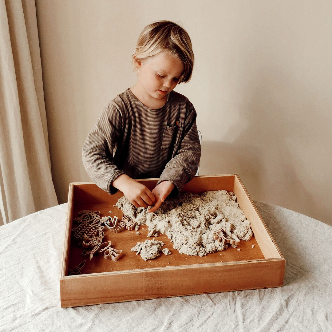 Een jongen speelt met beige kinetisch zand en dierenvormpjes in een houten bak.